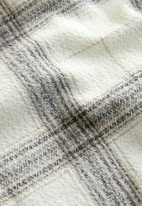 Tekstureeritud beež kangas, millel on tumedaid ja heledaid ruudumustreid, lähivaade, millelt on näha kootud niidid ja reljeefne tekstuur.