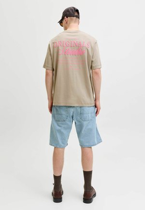 Mann iført beige T-skjorte med rosa "Originals Studio"-tekst på baksiden, lyseblå denimshorts, brune sokker, mørke sko, svart caps og solbriller.