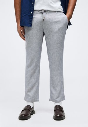 Personne portant un pantalon à cordon de serrage gris clair, des chaussettes blanches, des mocassins marron foncé, un t-shirt blanc et une chemise bleu foncé déboutonnée.