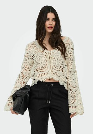 Femme aux longs cheveux bruns portant un chemisier en dentelle crochet crème et un pantalon noir à cordon, tenant une pochette noire pliée.