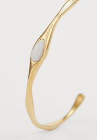 Pulsera de brazo de metal dorado con un diseño fluido y ondulado, y una piedra blanca ovalada lisa incrustada en el centro. Acabado pulido.