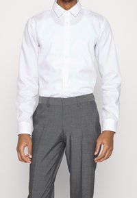 Biała koszula z długim rękawem i kołnierzykiem na guziki, zestawiona z szarymi, dopasowanymi spodniami o teksturowanej tkaninie i eleganckim kroju.