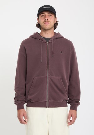 Homme portant un sweat à capuche zippé bordeaux avec poches avant et une casquette noire, debout devant un fond blanc uni.