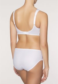 Vrouw in een witte bh en bijpassende hoge slip, van achteren afgebeeld tegen een neutrale lichte achtergrond.