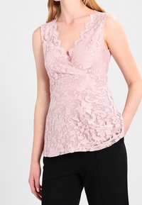 En rosa spetsblus med V-ringning, scallopade kanter och en figurnära silhuett, ihopparad med svarta byxor. Texturerad blommönster genom hela.