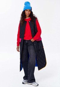 Žena v modré pletené čepici, červeném svetru, dlouhém černém prošívaném vestě s růžovými detaily, černých džínách a stříbrných teniskách, stojí se rukou v kapse.