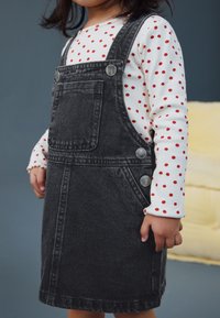 Robe salopette en denim gris avec poches latérales et boutons argentés, associée à un haut blanc à manches longues orné de pois rouges.