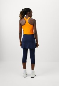 Narancssárga sportos ujjatlan felső racerback stílusban, sötétkék szoknyás leggings, textúrált anyag, és fehér sportcipő. A haj laza lófarokban van.