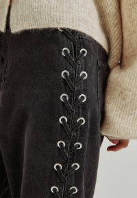 Pantalones de mezclilla negros con un diseño de cordones y ojales plateados a lo largo del lado, combinados con un suéter de punto beige claro.