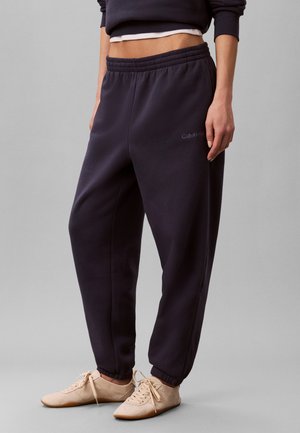 Person wearing dark navy sweatpants with elastic waist and cuffs, beige lace-up shoes, and cropped layered tops against a plain background.