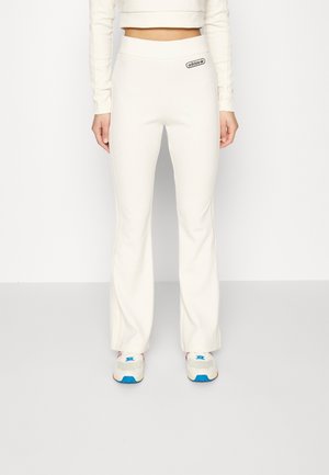 Femme portant un pantalon flare blanc Adidas, un top court manches longues et des baskets blanches avec des détails bleus et rouges.