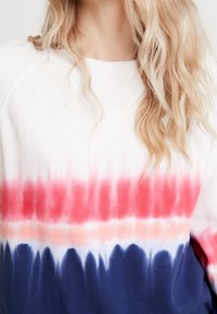 Personne blonde portant un sweat-shirt blanc avec des rayures tie-dye horizontales en rouge, rose clair et bleu foncé sur la poitrine et les manches.