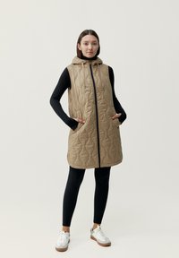 Beige vadderad väst med huva, framsida med dragkedja och sidofickor, bärs över en svart långärmad tröja och svarta leggings. Vita sneakers ingår.