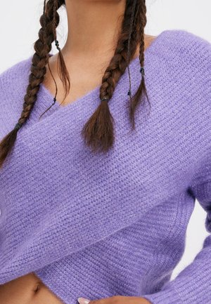 Gros plan d'une personne aux cheveux bruns tressés portant un pull lavande doux et texturé avec un décolleté en V croisé.