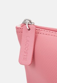 Ružičasta teksturirana torbica s srebrnim zatvaračem i ružičastom etiketom s utisnutim natpisom "LACOSTE". Glatki materijal, kompaktni pravokutni oblik.