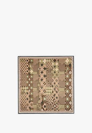 Femme aux cheveux bruns mi-longs porte un haut noir et une écharpe à motifs avec des formes géométriques dans des tons verts, bruns et beiges.