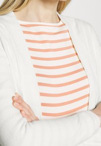 Cardigan blanco sobre una camisa de rayas blancas y naranja claro, con cuello redondo y textura de tela suave.