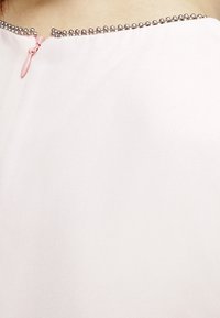 Vaaleanpunainen paita, jossa on tyylikäs viimeistely, pyöreä pääntie, jota korostaa rivissä olevat hopeiset helmet, sekä pieni vetoketjulukko takana.