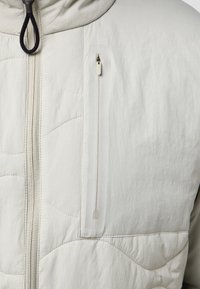 Veste isolante gris clair avec une poche poitrine zippée, un tissu texturé et une capuche avec un cordon de serrage. Les détails de couture sont visibles.