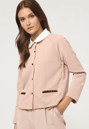 Chaqueta rosa texturizada con cuello blanco, botones negros y dos bolsillos frontales con ribete marrón oscuro. Presenta un corte relajado y mangas largas.
