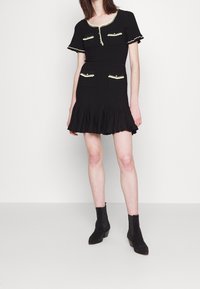 Schwarzes, geripptes Kleid mit kurzen Ärmeln, zwei vorderen Taschen und einem ausgestellten Rock. Mit kontrastierendem, hellem Besatz entlang des Ausschnitts und der Taschen.