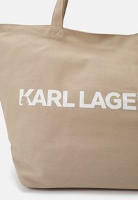 Beige canvas tote-väska med stadiga handtag, med vit tryckt text "KARL LAGERFELD." Slät textur och bred design för rymlig användning.