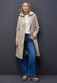 Beige Mantel mit Kapuze, Knopfverschlüssen und Seitentaschen, kombiniert mit einem weißen Kabelstrickpullover und blauen Schlagjeans. Braune Wildlederstiefel sind enthalten.