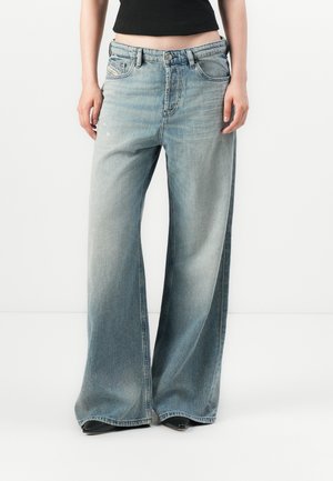 Persona che indossa jeans ampi di un leggero blu sbiadito e scarpe nere a punta, in piedi davanti a uno sfondo bianco uniforme.
