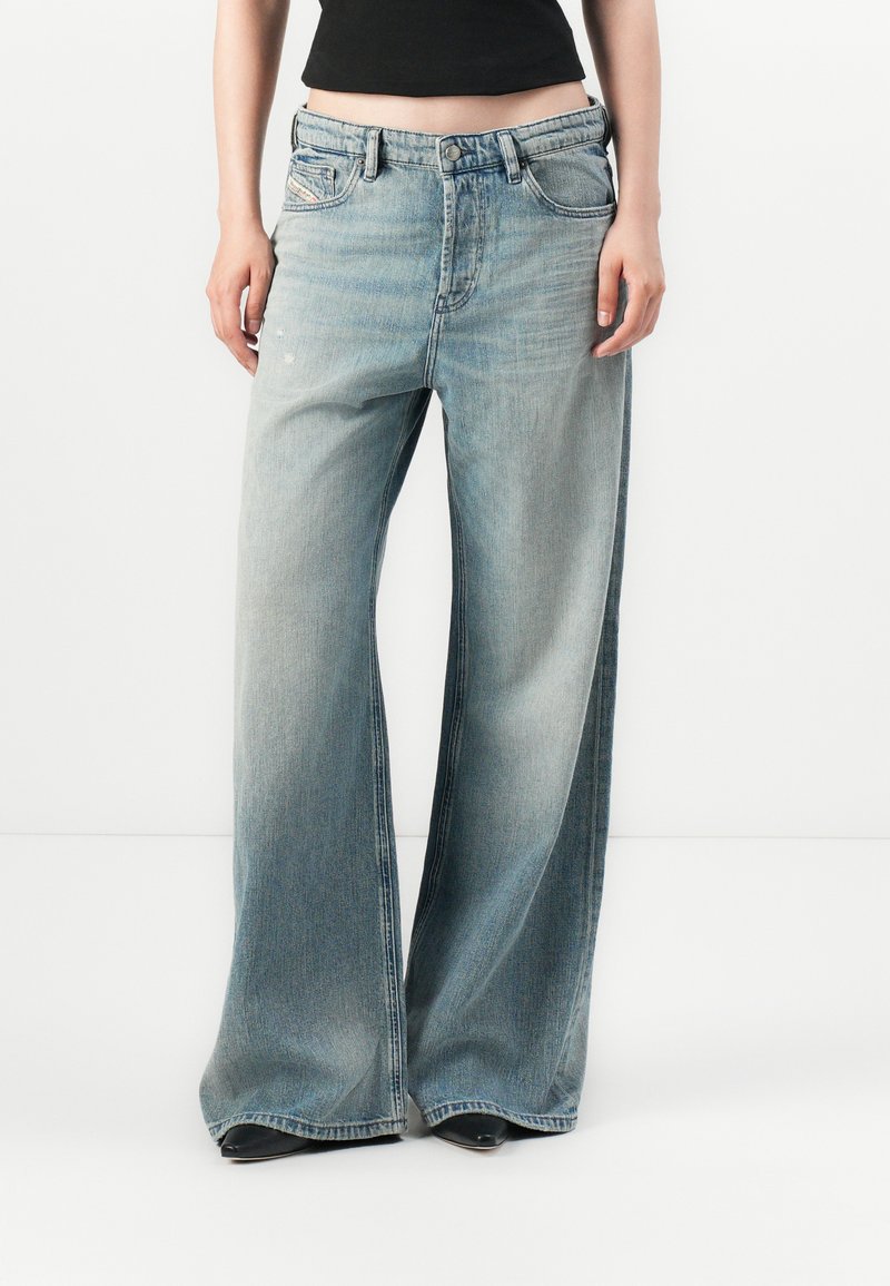 Person som har på seg lyseblå vid bukser med falmet denim og svarte spisse sko, står mot en vanlig hvit bakgrunn.