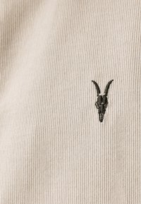 Tessuto di corduroy beige con righe verticali, dotato di un design ricamato di teschio di capra nero posizionato sul lato sinistro.