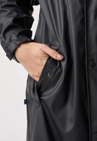 Czarny wodoodporny płaszcz o gładkiej fakturze, z boczną kieszenią i mankietami zapinanymi na guziki. Posiada małą niebiesko-żółtą metkę.