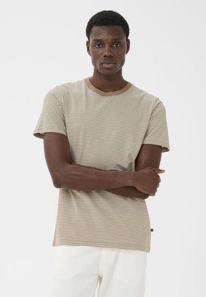 Ung mand står med armene krydsede iført en beige og hvidstribet t-shirt og hvide bukser mod en ensfarvet baggrund.