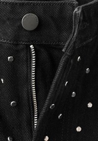 Acercamiento de unos jeans de mezclilla negra con cremallera plateada, botón de metal etiquetado "MNG JEANS 1984" y pequeños remaches plateados en la tela.