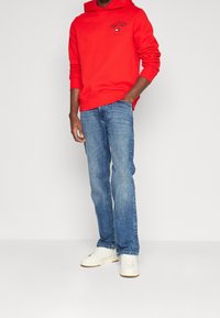 Rode hoodie met een grafisch ontwerp aan de voorkant, gecombineerd met blauwe rechte jeans en witte sneakers, tegen een neutrale achtergrond.