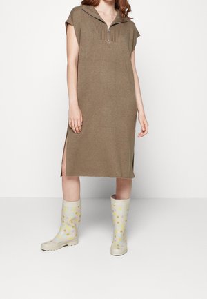 Nainen pukeutuneena polvipituiseen ruskeaan neulemekkoon, jossa on lyhyet hihat, sivuhalkiot ja beiget kumisaappaat, joissa on värikkäitä pilkkuja.