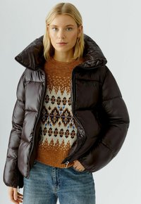 Dunkelbrauner Puffer-Jacke mit hohem Kragen und Reißverschluss, kombiniert mit einem gemusterten Strickpullover in warmen Tönen, getragen mit blauen Jeans.