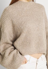 Beige stickad tröja med hög hals, vida ärmar och ribbad textur. Avskuren design som visar midjan, kombinerad med vita jeans.