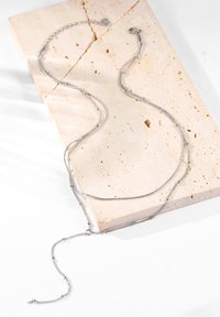 Srebrny naszyjnik łańcuszkowy z wieloma pasmami i różnymi ogniwami, umieszczony na teksturowanej beżowej powierzchni kamienia. Prosty i nowoczesny design.