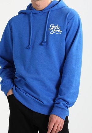 Mann trägt eine blaue Jack & Jones Kapuzenjacke mit Kordelzug, schwarze Hose und hat die Hand in der Tasche vor einem schlichten hellen Hintergrund.