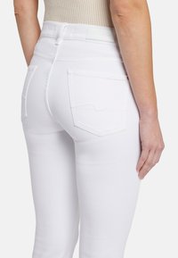 Jean blanc taille haute avec une texture lisse, dotée de deux poches arrière et de détails de couture subtils. La main du modèle repose sur la hanche.