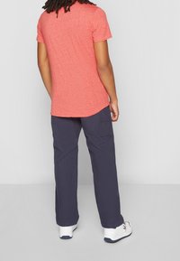 Persona vistiendo una camisa de manga corta color coral, pantalones de carga color navy y zapatillas blancas, de pie de espaldas contra un fondo gris claro liso.