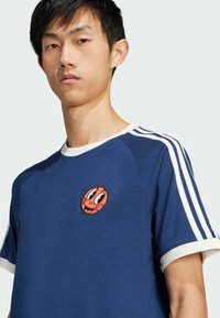 Navy blue t-shirt with white stripes on sleeves, featuring a round orange patch with a cartoon face at the chest. Soft fabric texture.