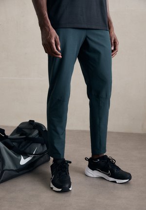 Dark green athletic pants with a tapered design, paired with black sneakers and a gray duffel bag featuring a white logo.