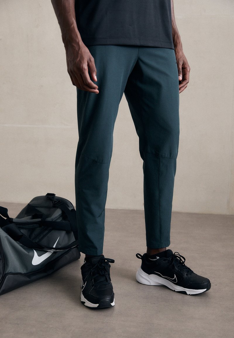 Mørkegrønne træningsbukser med en tapered design, parret med sorte sneakers og en grå duffeltaske med et hvidt logo.