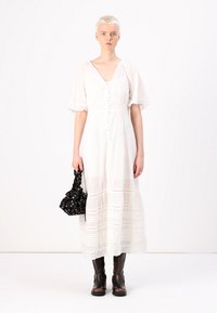 Robe maxi blanche fluide avec des manches courtes bouffantes, un décolleté boutonné et un ourlet à volants. Le mannequin tient un sac à main noir à motifs floraux et porte des bottes marron.