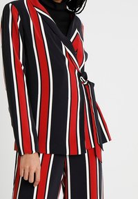 Veste à rayures noire, rouge et blanche, avec une coupe ajustée, des manches longues et un détail de ceinture nouée à la taille.