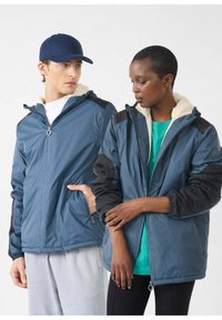 Chaquetas con cremallera en azul y negro con forro polar, con bolsillos y puños elásticos, usadas por dos personas sobre un fondo blanco.