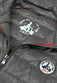 Giacca trapuntata nera con cerniera argentata, con logo di un pilota di scooter bianco e testo "Just Over The Top JOTT" sulla parte anteriore e sulla fodera interna.