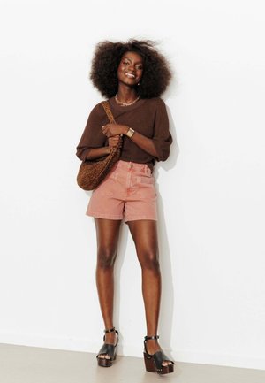 Femme souriante aux cheveux bouclés et volumineux, portant un pull marron, un short rose, des sandales compensées noires et un sac à bandoulière imprimé léopard.