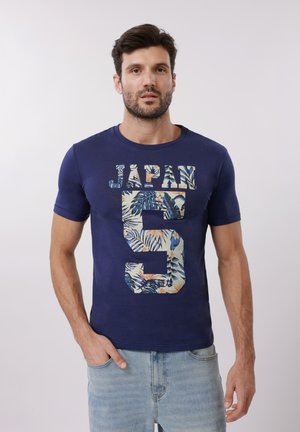 Mann trägt ein dunkelblaues T-Shirt mit der Aufschrift "JAPAN 5" und Blumenmuster, kombiniert mit hellblauen Jeans, und steht vor einem einfarbigen Hintergrund.